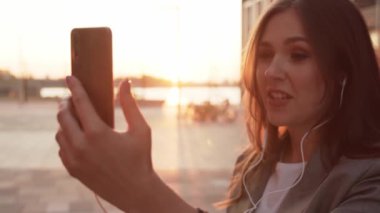 Dışarıda bankta oturan çekici genç bir iş kadını. Video kaset için akıllı telefon kullanan bir kız. Sosyal uzaklık kavramı. Gün batımı ışığı.