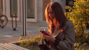 Genç çekici iş kadını dışarıda bankta oturuyor ve akıllı telefon kullanıyor. Gün batımı ışığı.