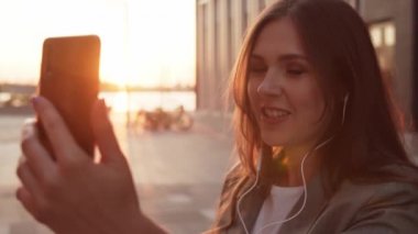 Dışarıda bankta oturan çekici genç bir iş kadını. Video kaset için akıllı telefon kullanan bir kız. Sosyal uzaklık kavramı. Gün batımı ışığı.