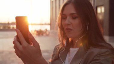 Dışarıda bankta oturan çekici genç bir iş kadını. Video kaset için akıllı telefon kullanan bir kız. Sosyal uzaklık kavramı. Gün batımı ışığı.