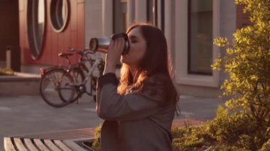 Genç ve çekici bir iş kadını bankta oturuyor ve kahve içiyor. Gün batımı ışığı.