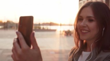 Dışarıda bankta oturan çekici genç bir iş kadını. Video kaset için akıllı telefon kullanan bir kız. Sosyal uzaklık kavramı. Gün batımı ışığı.
