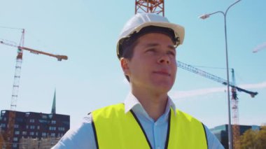 İnşaat alanının önünde duran profesyonel müteahhit. Kasklı ve yelekli ustabaşı. Ofis binası ve vinç arka planı. İş, gayrimenkul ve yatırım kavramları.