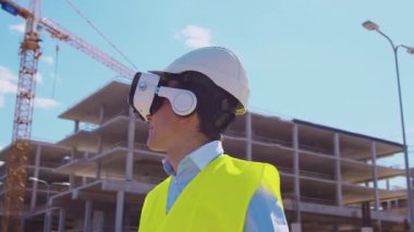 İnşaat alanının önünde sanal gerçeklik teknolojileri kullanarak VR kasklı profesyonel bir inşaatçı. Ofis binası ve vinç arka planı. Emlak ve yatırım kavramları