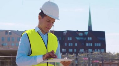 İnşaat alanının önünde elinde tablet bilgisayarla duran profesyonel müteahhit. Miğferli ve yelekli ustabaşı. Ofis binası ve vinç arka planı. İş, gayrimenkul ve yatırım kavramları.