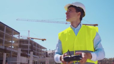 Drone operatörü uzaktan kumanda tutuyor. İnşaat alanının önünde miğfer ve yelek giymiş profesyonel bir müteahhit duruyor. Ofis binası ve vinç arka planı. İş, gayrimenkul ve yatırım