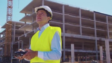 Drone operatörü uzaktan kumanda tutuyor. İnşaat alanının önünde miğfer ve yelek giymiş profesyonel bir müteahhit duruyor. Ofis binası ve vinç arka planı. İş, gayrimenkul ve yatırım