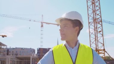 İnşaat alanının önünde duran profesyonel müteahhit. Kasklı ve yelekli ustabaşı. Ofis binası ve vinç arka planı. İş, gayrimenkul ve yatırım kavramları.