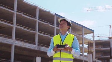 Drone operatörü uzaktan kumanda tutuyor. İnşaat alanının önünde miğfer ve yelek giymiş profesyonel bir müteahhit duruyor. Ofis binası ve vinç arka planı. İş, gayrimenkul ve yatırım
