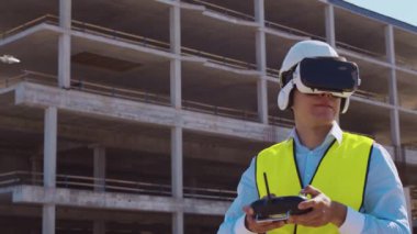 Sanal gerçeklik kasklı profesyonel dron operatörü inşaat alanının önünde duruyor. Uzaktan kumandalı bir bina. Ofis binası ve vinç arka planı. İş, gayrimenkul ve