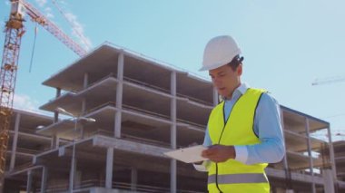 İnşaat alanının önünde elinde tablet bilgisayarla duran profesyonel müteahhit. Miğferli ve yelekli ustabaşı. Ofis binası ve vinç arka planı. İş, gayrimenkul ve yatırım kavramları.