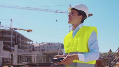 Drone operatörü uzaktan kumanda tutuyor. İnşaat alanının önünde miğfer ve yelek giymiş profesyonel bir müteahhit duruyor. Ofis binası ve vinç arka planı. İş, gayrimenkul ve yatırım