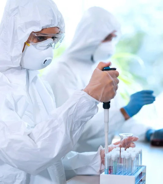 Scientists in protection suits and masks working in research lab using laboratory equipment: microscopes, test tubes. Biological hazard, pharmaceutical discovery, bacteriology and virology concept.