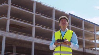 Drone operatörü uzaktan kumanda tutuyor. İnşaat alanının önünde miğfer ve yelek giymiş profesyonel bir müteahhit duruyor. Ofis binası ve vinç arka planı. İş, gayrimenkul ve yatırım