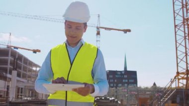 İnşaat alanının önünde elinde tablet bilgisayarla duran profesyonel müteahhit. Miğferli ve yelekli ustabaşı. Ofis binası ve vinç arka planı. İş, gayrimenkul ve yatırım kavramları.