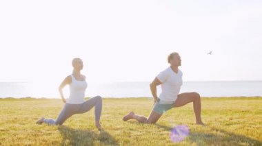 Formda bir adam ve güzel bir kadın çimlerin üzerinde yoga yapıyor. Deniz, gökyüzü ve arka plandaki ağaç. Gün batımında esneme egzersizi. Spor, spor, sağlık hizmetleri ve yaşam tarzı kavramları.