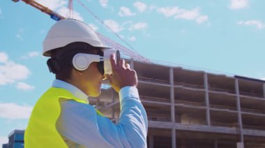 İnşaat alanının önünde sanal gerçeklik teknolojileri kullanarak VR kasklı profesyonel bir inşaatçı. Ofis binası ve vinç arka planı. Emlak ve yatırım kavramları