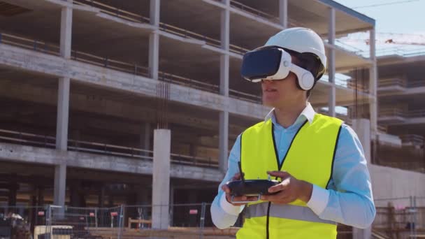 专业的无人机操作员戴着虚拟现实头盔站在建筑工地前 建设者持有远程控制器 办公大楼和起重机背景 房地产和 — 图库视频影像
