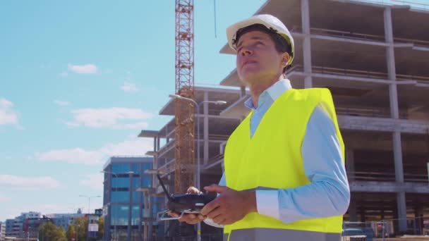 无人机操作员拿着遥控器身穿头盔和背心的专业建筑商站在建筑工地前 办公大楼和起重机背景 房地产和投资 — 图库视频影像