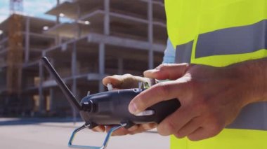 Drone operatörü uzaktan kumanda tutuyor. İnşaat alanının önünde miğfer ve yelek giymiş profesyonel bir müteahhit duruyor. Ofis binası ve vinç arka planı. İş, gayrimenkul ve yatırım
