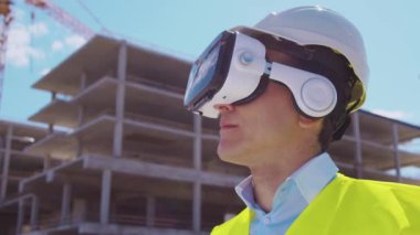 Sanal gerçeklik kasklı profesyonel dron operatörü inşaat alanının önünde duruyor. Uzaktan kumandalı bir bina. Ofis binası ve vinç arka planı. İş, gayrimenkul ve