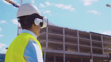 İnşaat alanının önünde sanal gerçeklik teknolojileri kullanarak VR kasklı profesyonel bir inşaatçı. Ofis binası ve vinç arka planı. Emlak ve yatırım kavramları