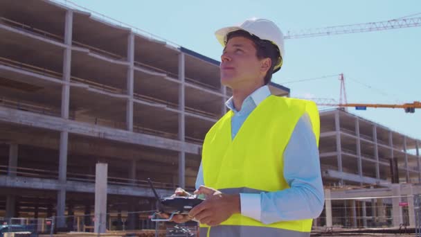 无人机操作员拿着遥控器身穿头盔和背心的专业建筑商站在建筑工地前 办公大楼和起重机背景 房地产和投资 — 图库视频影像
