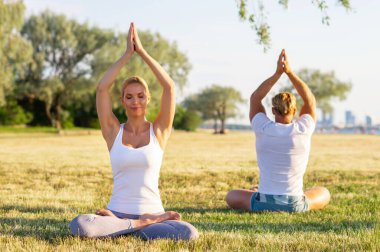 Formda bir adam ve güzel bir kadın çimlerin üzerinde yoga yapıyor. Arka planda deniz ve gökyüzü var. Gün batımında esneme egzersizi. Spor, spor, sağlık hizmetleri ve yaşam tarzı kavramları.