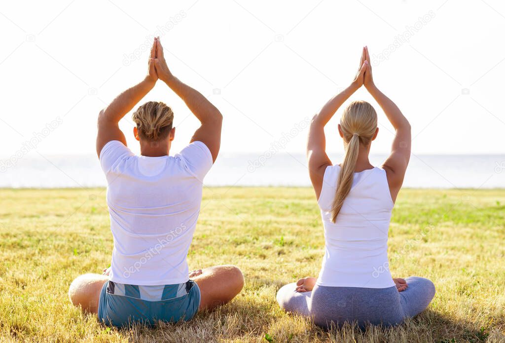 Fit man and beautiful woman practicing yoga outdoor on the grass. Sea and sky on the background. Stretching exercise in the sunset. Sport, fitness, health care and lifestyle concepts.