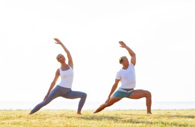 Formda bir adam ve güzel bir kadın çimlerin üzerinde yoga yapıyor. Arka planda deniz ve gökyüzü var. Gün batımında esneme egzersizi. Spor, spor, sağlık hizmetleri ve yaşam tarzı kavramları.