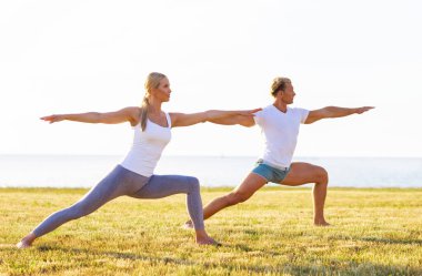 Formda bir adam ve güzel bir kadın çimlerin üzerinde yoga yapıyor. Arka planda deniz ve gökyüzü var. Gün batımında esneme egzersizi. Spor, spor, sağlık hizmetleri ve yaşam tarzı kavramları.
