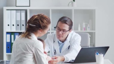 Erkek çocuk doktoru küçük hasta bebeği muayene ediyor. Doktor, anne ve kızı klinikteki muayenehanede..