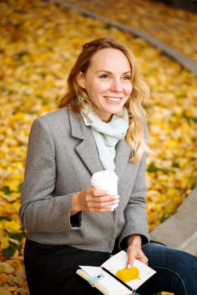 Gelukkig blond glimlachend vrouw met papieren notitieboekje en koffie drinken — Stockfoto