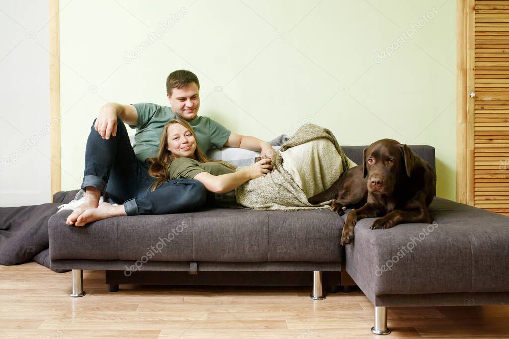 Young family couple in casual clothes with dog labrador retriever, woman lying on man`s knees, stay home concept, full body