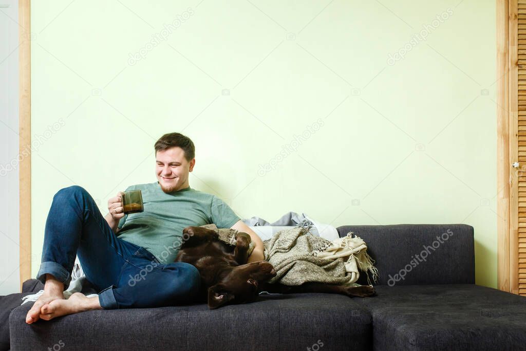 Happy handsome man sitting on the couch at home with pet dog labrador retriever lying on his knees, dog canine therapy concept