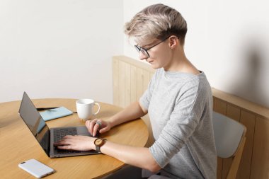 Genç odaklı hippi milenyum kadın, kısa sarı saç kesimi, laptopla kafenin içinde çalışıyor, yakın çekimde, ekrana bakıyor ve daktilo ediyor.