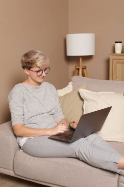 Genç mutlu kadın serbest çalışan ya da evden öğrenen. 30 'lu yaşlarda, rahat kıyafetler ve gözlüklerle kanepede oturmuş dizüstü bilgisayarla gülümseyen oldukça zeki bir kadının vücut portresi..