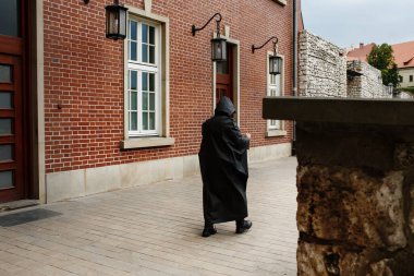 Koyu siyah yağmurluklu tanınmayan gizemli adam Avrupa 'nın eski bir şehrinde kırmızı tuğla duvar boyunca yürüyor.