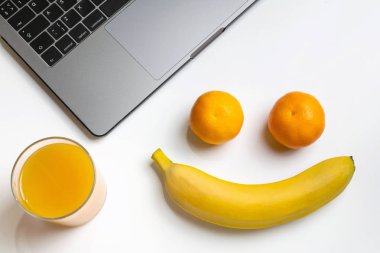 Çalışma alanı üzerinde meyve. Komik gülümsemek yüz. Beyaz masa üzerinde laptop, muz, mandalina ve portakal suyu. Meyve çalışmaları ile snack bir lokma.