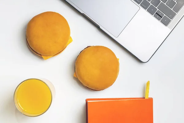 Fast food at work snacking. Laptop, notebook, two hamburgers and french fry at workplace. Snacking on white desk.