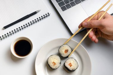 Mola zamanı suşi yemek için. İş yerinde yemek suşi rulo. Laptop ve defter beyaz organize masa üzerinde.