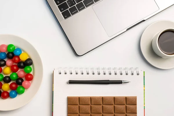 Snacking at work, have a bite concept. Laptop, candy and workbook with bar of chocolate and coffee at workplace. Flat lay, top view, white table.