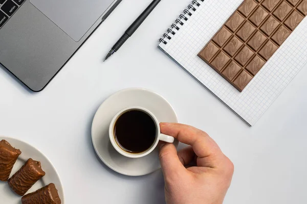 Snacking at work, have a bite concept. Laptop, candy and workbook with bar of chocolate. Men hand with cup of coffee at workplace. Flat lay, top view, white table.