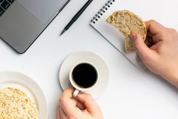 Healthy snacking at work during break time. Crispy rice rounds with coffee near the laptop and notebook. White organized desk.