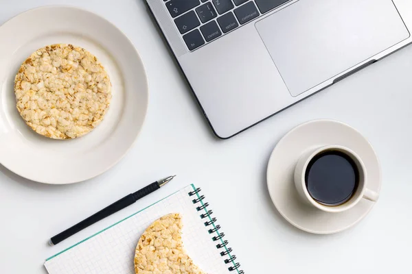Healthy snacking at work during break time. Crispy rice rounds with coffee near the laptop and notebook. White organized desk.