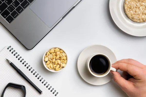 Healthy snacking at work during break time. Sportsman eating crispy rice rounds with peanuts, cup of coffee near the laptop, fitness-tracker and notebook. White organized desk.