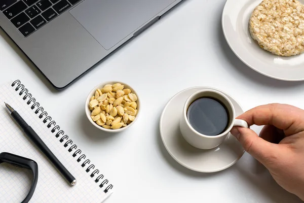 Healthy snacking at work during break time. Sportsman eating crispy rice rounds with peanuts, cup of coffee near the laptop, fitness-tracker and notebook. White organized desk.