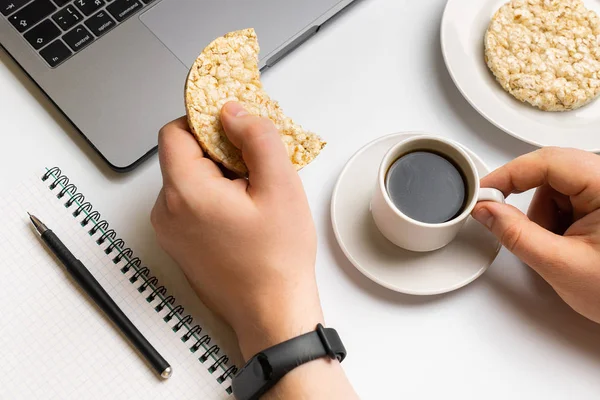 Healthy snacking at work during break time. Sportsman eating crispy rice rounds with coffee near the laptop and notebook. White organized desk.