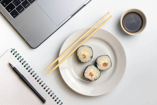 Sushi rolls snacking at work. Break time for sushi eating. Laptop and notebook on white organized desk.