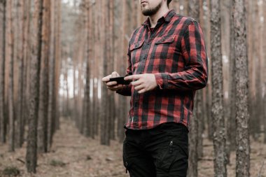 Kırmızı ekose gömlek ve siyah askeri pantolon adam elinde bir smartphone tutar. Çam ormanında kaybolmuş. Boşluk Kopyala. Tonda